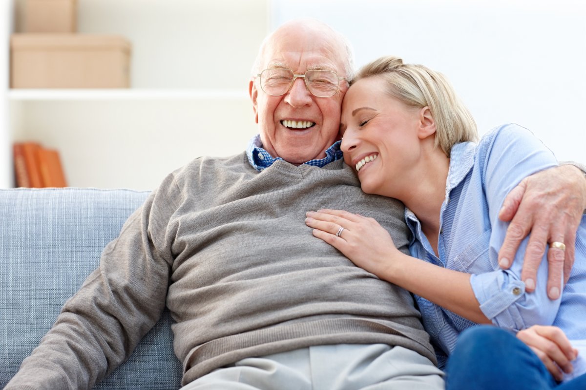 An elderly man in gray laughs with his daughter on the couch