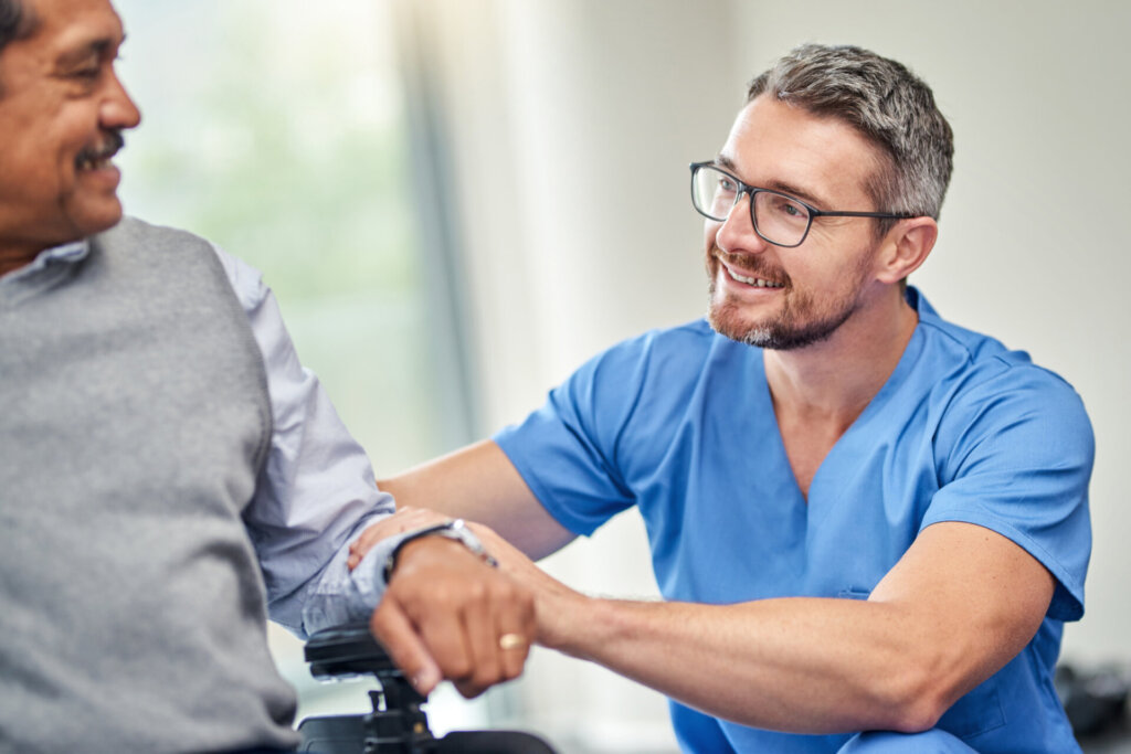 A male memory care, senior living resident sitting in a wheelchair, looking at a male caregiver wearing blue scrubs kneeling beside him. Traditions Meadow Brook Senior Living in Fishers, IN.