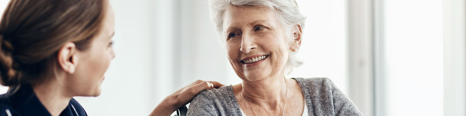 A senior woman with gray hair sitting in a wheelchair smiling and looking over at her female assisted living nurse