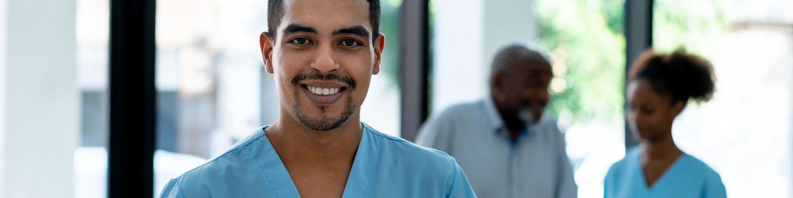 A male assisted living nurse wearing blue scrubs and smiling as a female assisted living nurse helps a male senior patient in the background