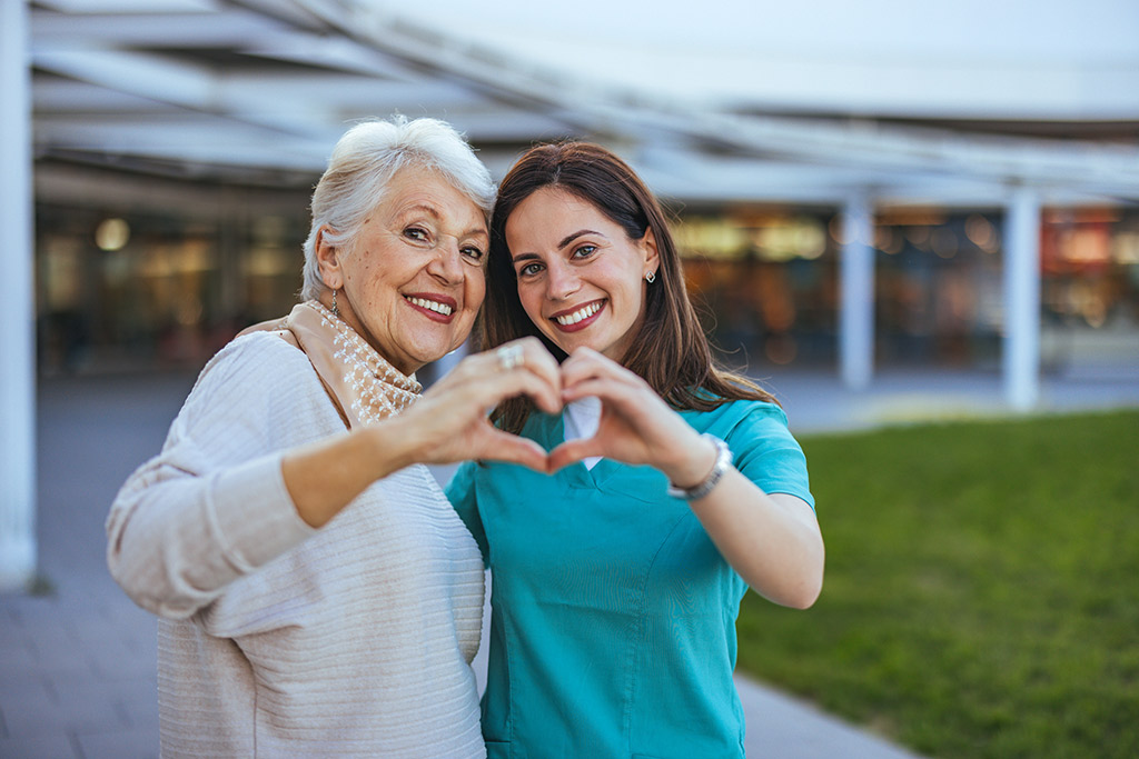 senior woman in grey shirt standing with younger woman in blue scrub top holding their hands together in a heart